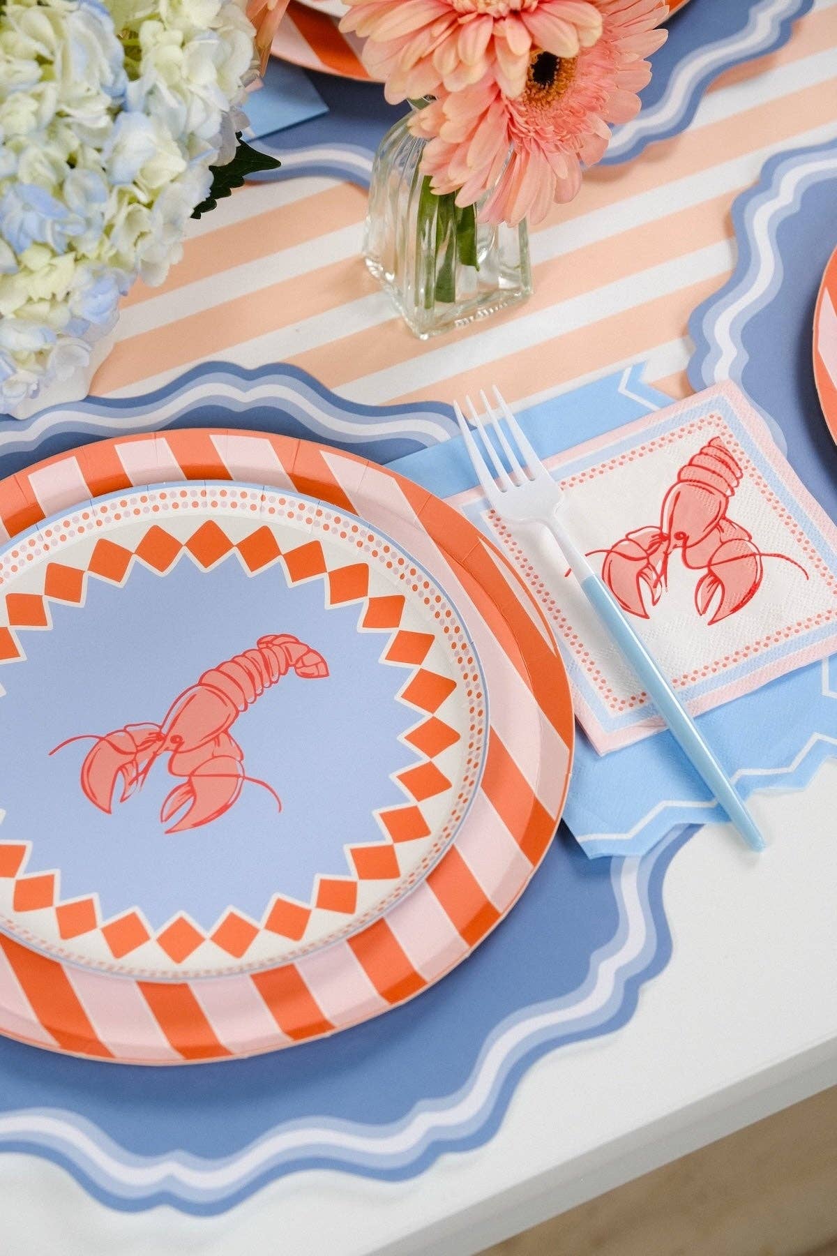  Close-up angled shot of a single place setting on a blue double-wave scalloped placemat with a peach-stripe table runner behind. The lobster salad plate sits over an orange and pink diagonal stripe charger plate. Blue plastic fork and knife resting on the plate. Lobster cocktail napkin with pink and blue border positioned to the right. Peach gerbera daisy in a glass bud vase in the background.