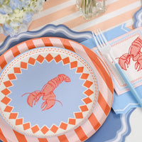 Straight-on angled view of one lobster place setting on the blue-and-white stripe runner. Orange stripe charger below, lobster salad plate on top with blue fork and knife in place. Lobster napkins visible on both left and right sides of the place setting. Tall peach gerbera and white daisy arrangements on both sides.