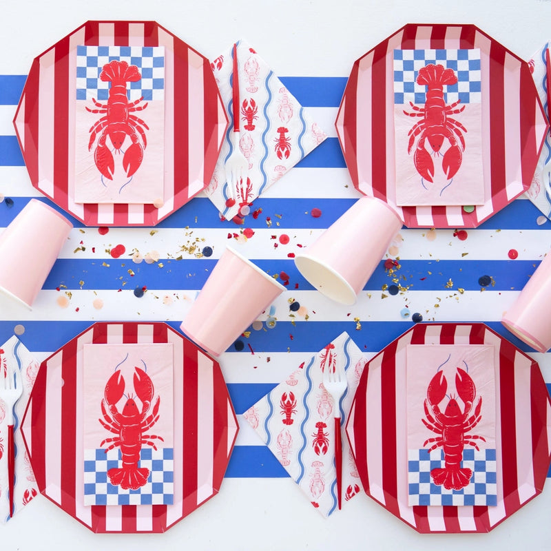 Overhead view of nautical tablescape featuring red‑striped plates, lobster guest towels and pastel cups on blue‑and‑white runner with confetti.