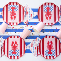 Nautical tablescape with striped red plates, lobster lunch napkins, pink cups and blue‑white runner scattered with confetti.