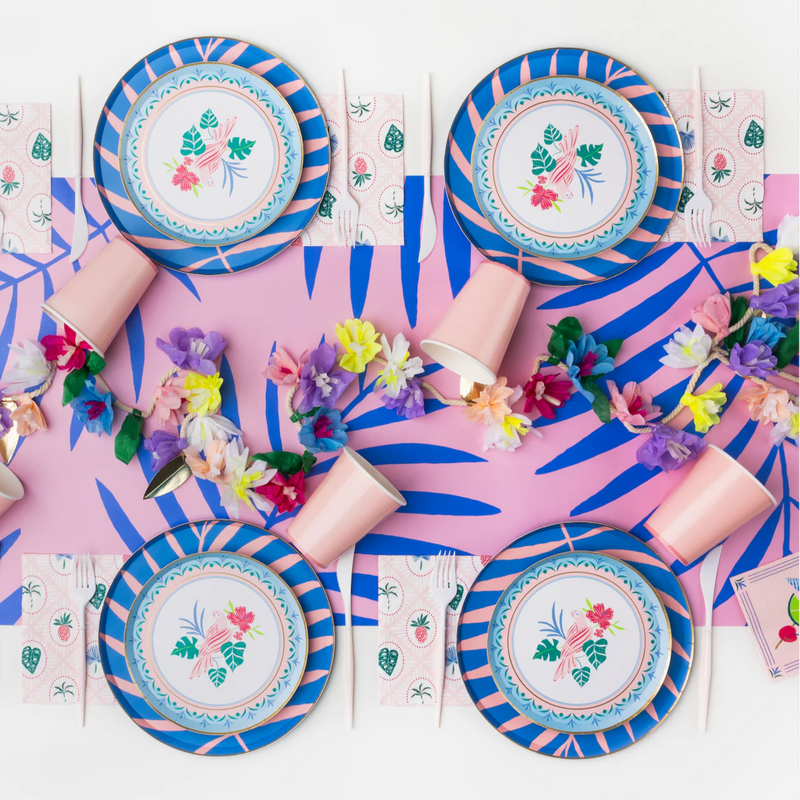 Pink and blue palm‑leaf table runner under tropical plates, pink cups, and lei garland at a luau setup.