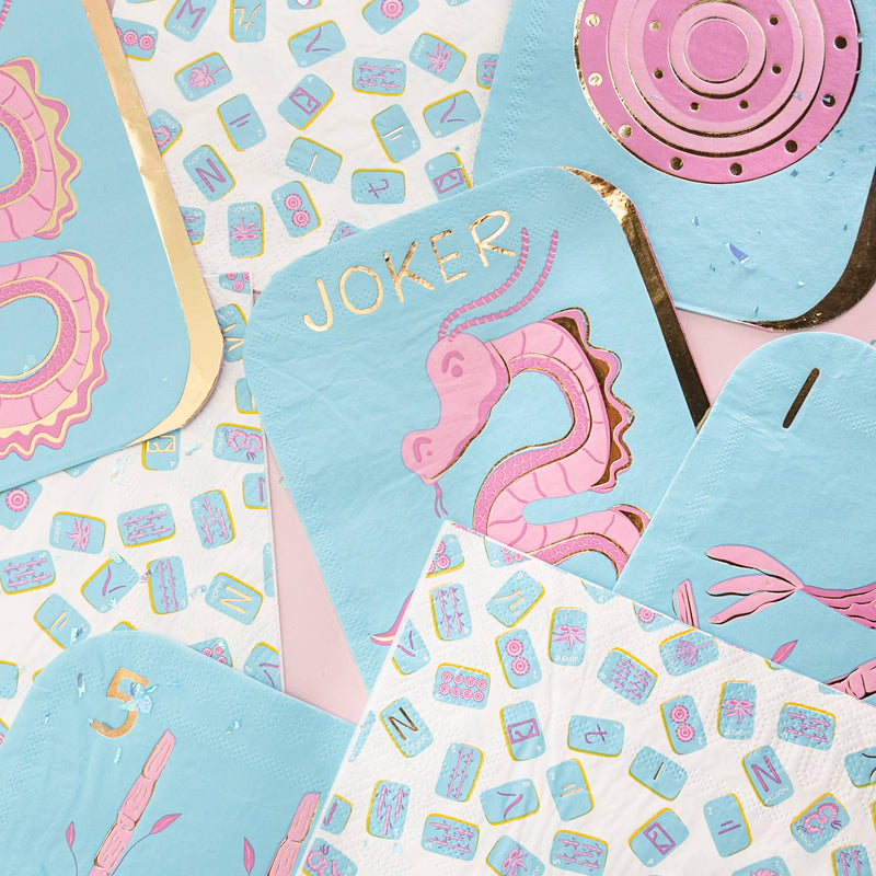 Flatlay of mahjong tile shaped paper napkins in turquoise with pink and gold foil designs, scattered with all-over mahjong tile print napkins and pink dot tile shaped napkins, showing Joker tile design with gold foil "JOKER" lettering and pink dragon illustration