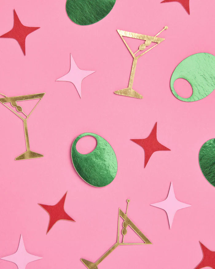 Assorted martini party confetti pieces including gold martini glasses, green olives, and pink and red stars scattered on pink background