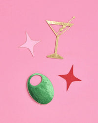 Single gold foil martini glass confetti piece with green olive, pink star, and red star arranged on pink background