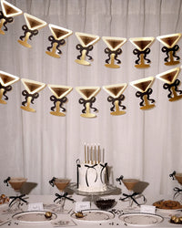 Two rows of martini glass bow banners hanging above an espresso martini birthday table with cake, chocolates, place cards, and cocktails