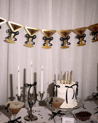 Martini glass bow banner hanging above espresso martini birthday dessert table with candlelit cake and cocktails
