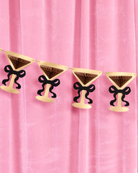Gold foil martini glass bow banner hung against pink velvet backdrop with brown tissue accordions and black paper bows