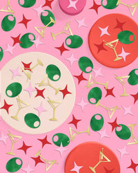 Gold martini glass confetti with green olives and pink and red stars scattered across pink background with cocktail-themed paper plates