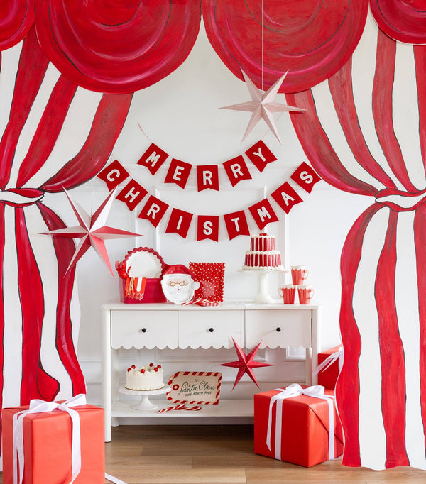 The Merry Christmas felt banner strung on a mantel against a red-and-white striped backdrop with red balloons.