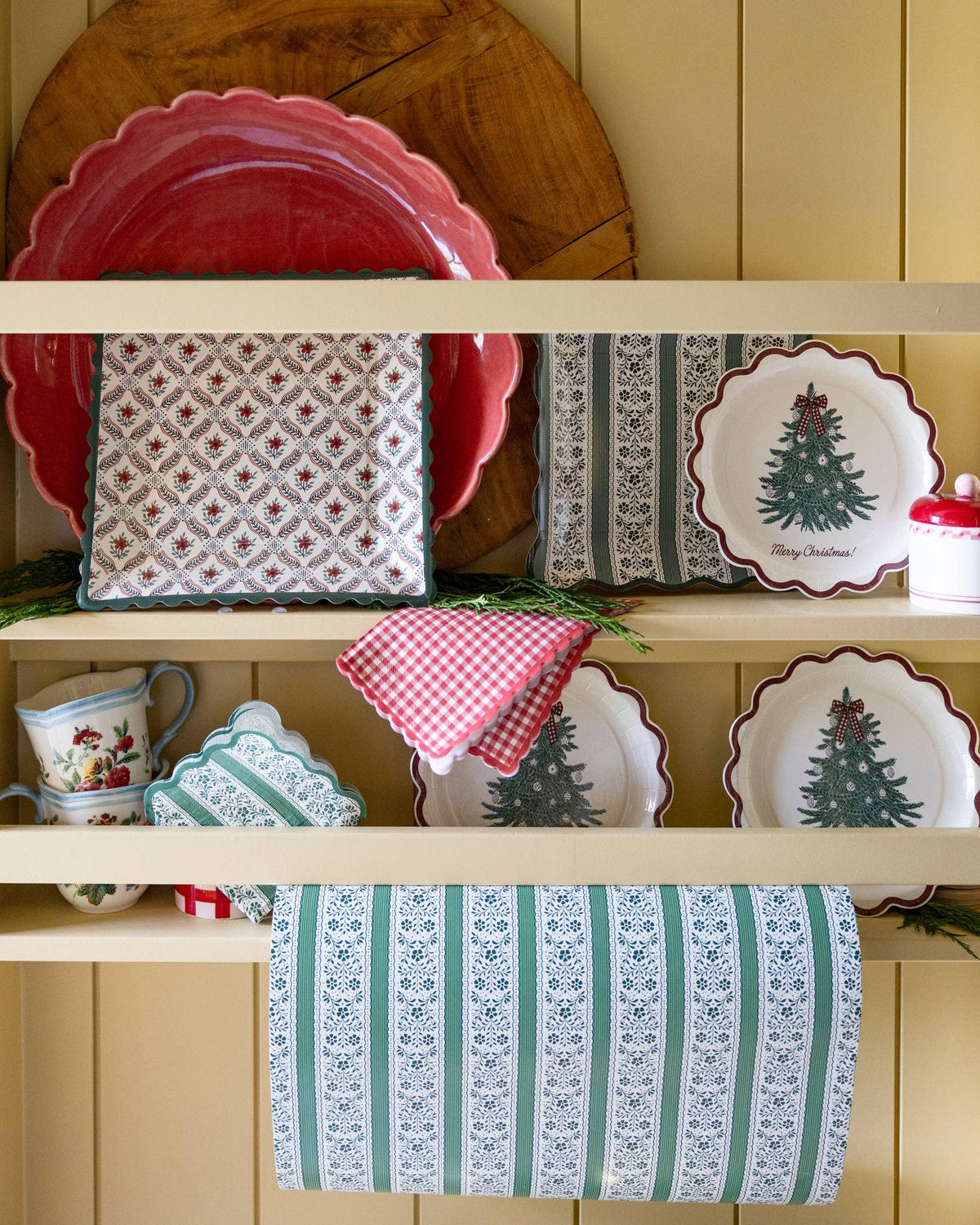 Merry Christmas tree scalloped paper plates displayed on a cream kitchen hutch with red gingham napkins, printed paper goods, and fresh cedar greenery