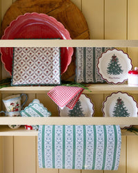 Merry Christmas tree scalloped paper plates displayed on a cream kitchen hutch with red gingham napkins, printed paper goods, and fresh cedar greenery