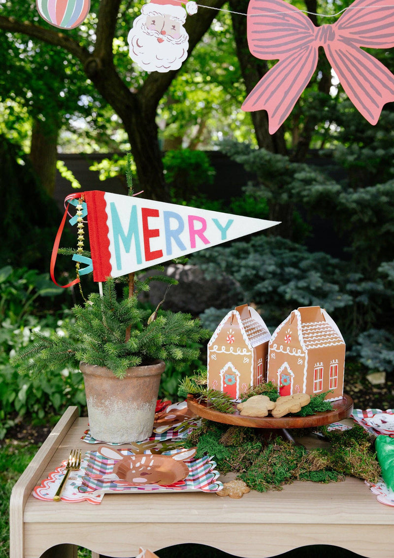 MERRY felt pennant in a potted spruce on an outdoor party table; gingerbread house treat boxes, plates, and garland shown separately, not included