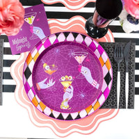 Overhead Halloween place setting with a purple Midnight Spirits plate showing the eyeball martini and hand design, pink scalloped edge, black diamond border, and black flatware