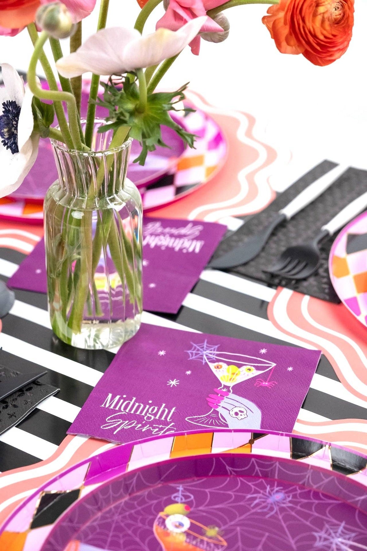 Halloween table setting with purple Midnight Spirits plates, a glass vase of pink and orange flowers, and a black and white striped runner