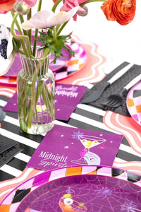 Halloween table setting with purple Midnight Spirits plates, a glass vase of pink and orange flowers, and a black and white striped runner