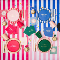Overhead lifestyle shot of colorful milestone birthday party table with assorted Happy Birthday plates, napkins and cups in pink, blue and green on striped tablecloths with confetti