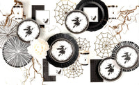Flat lay of scattered black cat cocktail napkins, vintage witch dessert plates, graphic spiderwebs, and white florals on a white surface.