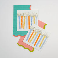 Two square beverage napkins with tall pastel candles and gold starbursts layered on teal and pink scalloped towels.