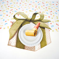 Place setting styled on a confetti table runner with a painted gift box and green bow under a white plate and party blower.