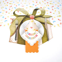 Party hat plate and silverware arranged on a confetti table runner for a birthday setting.
