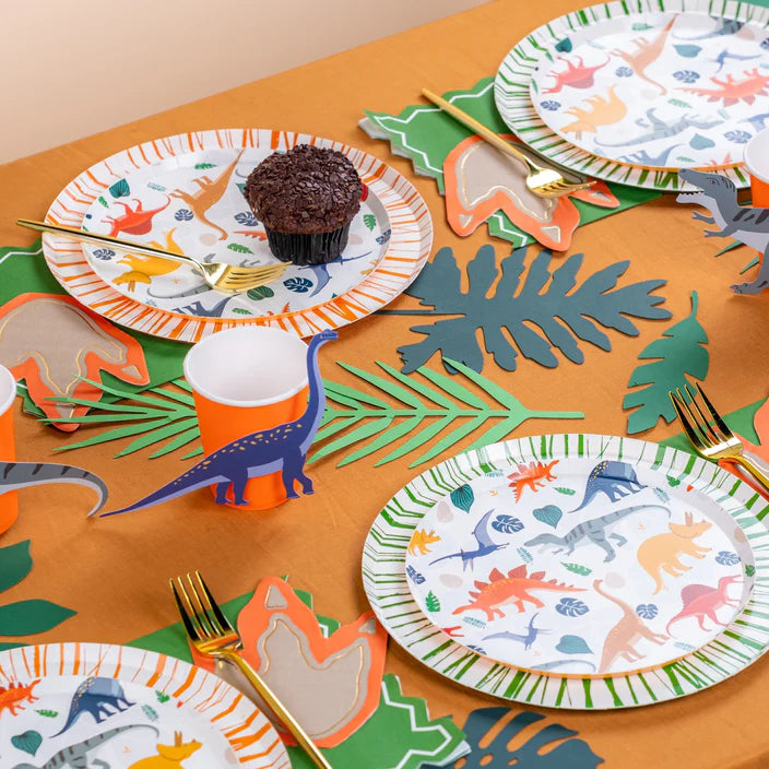 Dino party table with jungle leaves, orange cups, and dinosaur dessert plates.