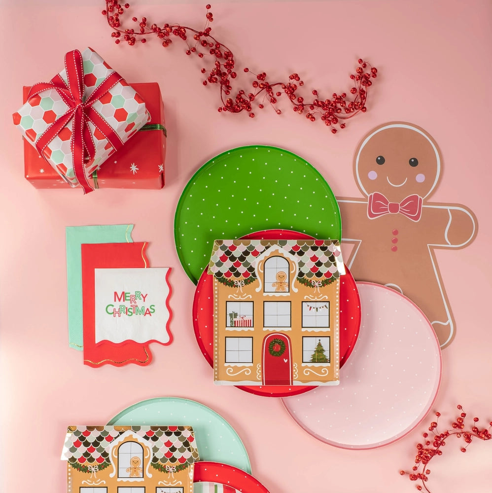 Flat lay of holiday plates, napkins, and a gingerbread placemat on a pink backdrop.