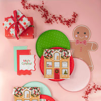 Flat lay of holiday plates, napkins, and a gingerbread placemat on a pink backdrop.