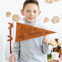 Child in a gray sweater holding a brown Give Thanks felt pennant in a decorated fall room.