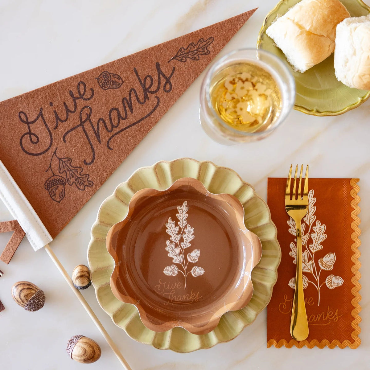 Flat lay with Give Thanks felt pennant beside scalloped fall plates, acorns, and a drink glass.