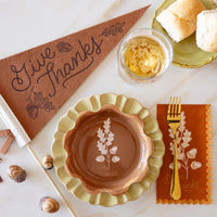 Flat lay with Give Thanks felt pennant beside scalloped fall plates, acorns, and a drink glass.