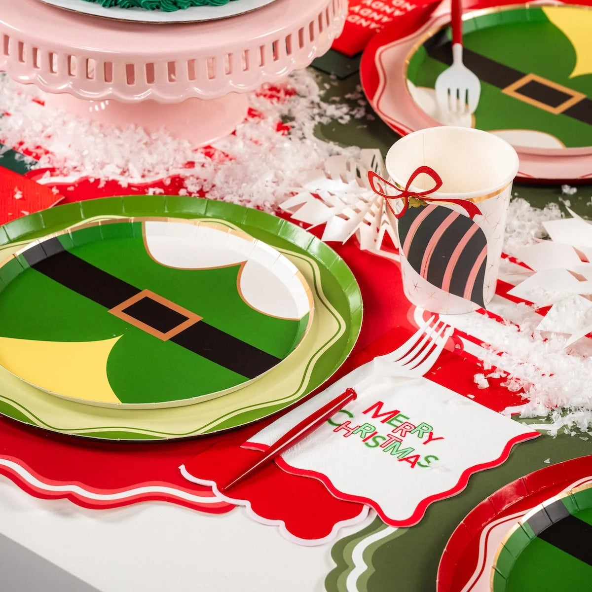 Pink and green cake on festive table beside elf plates and Merry Christmas napkin.