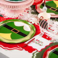 Pink and green cake on festive table beside elf plates and Merry Christmas napkin.