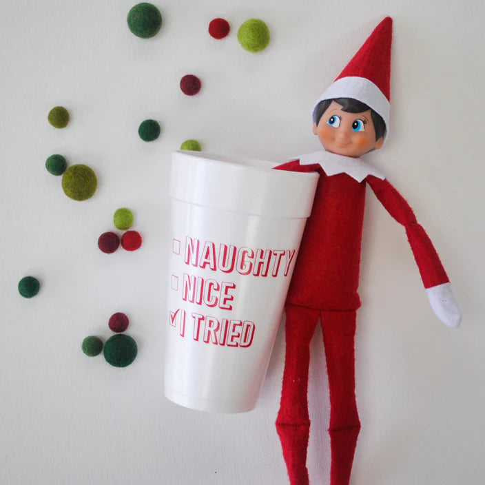 Red holiday elf doll holding a white 20 ounce foam cup with a red Naughty, Nice, checked I Tried checklist, with green and red felt balls nearby.