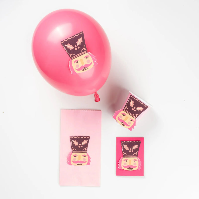 Pink balloon and paper cup decorated with nutcracker accent stickers next to a treat bag.