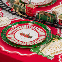 Place setting with train dessert plate layered on green argyle dinner plate beside toy tracks.