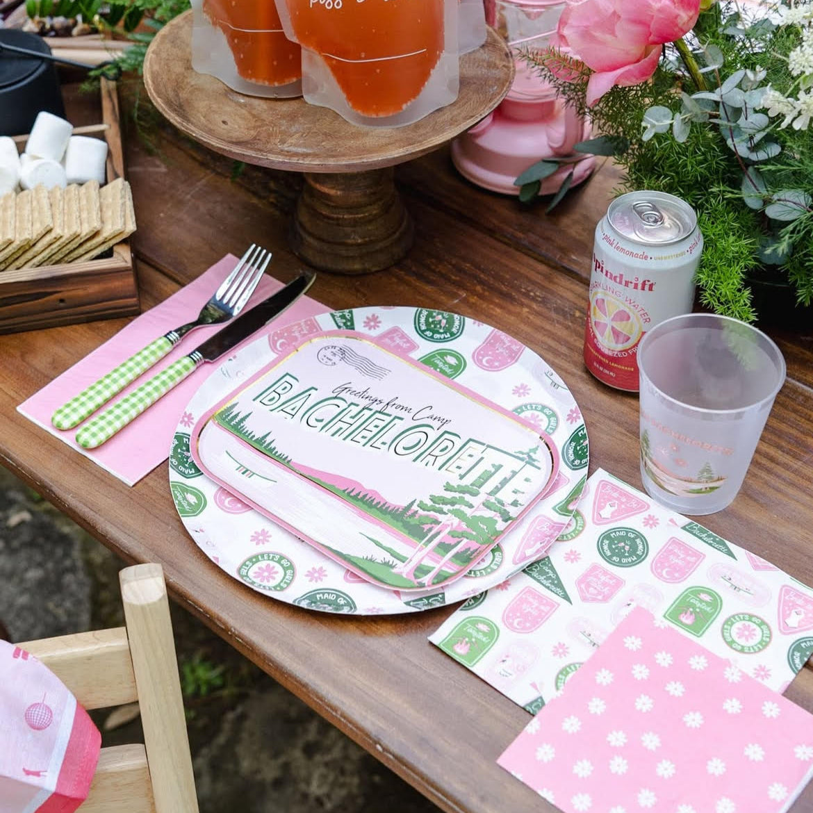 Rustic picnic table set with Camp Bachelorette badge dinner plate, matching napkins, pink drinks and s’mores fixings