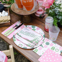 Rustic picnic table set with Camp Bachelorette plate, napkins, and flex cup beside pink flowers