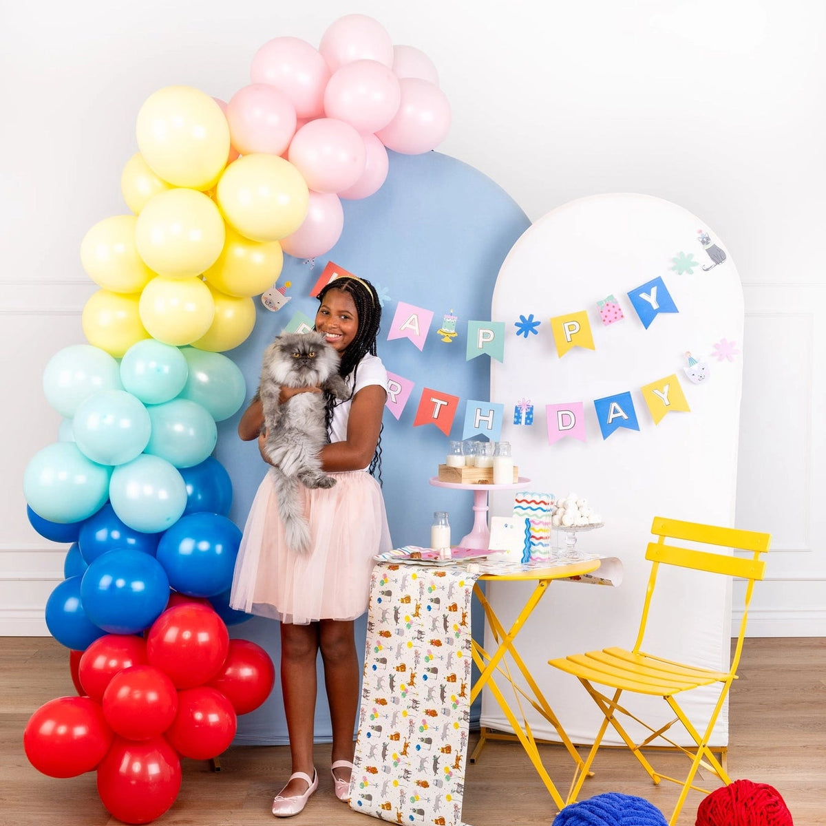 Girl holding fluffy cat beside pastel Cat Happy Birthday Banner and rainbow balloon arch