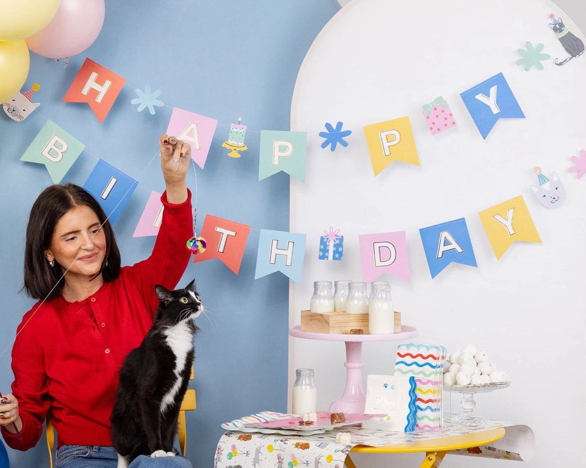 Woman playing with black-and-white cat under pastel Cat Happy Birthday Banner and balloons