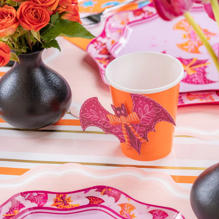 Close view of orange cup with pink bat sticker beside black vase and plates on a Halloween table.