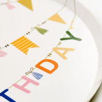 Close up of Happy Birthday bunting with tassels and flags on a white dessert plate.