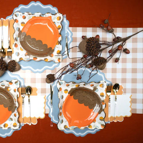 Thanksgiving table set on a brown gingham runner with orange plates, blue scalloped placemats, acorn accents, and branches.