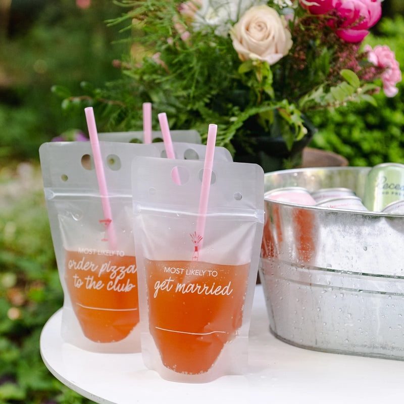 Three clear zip-top bachelorette drink pouches with pink straws on a garden bar next to a metal ice bucket.