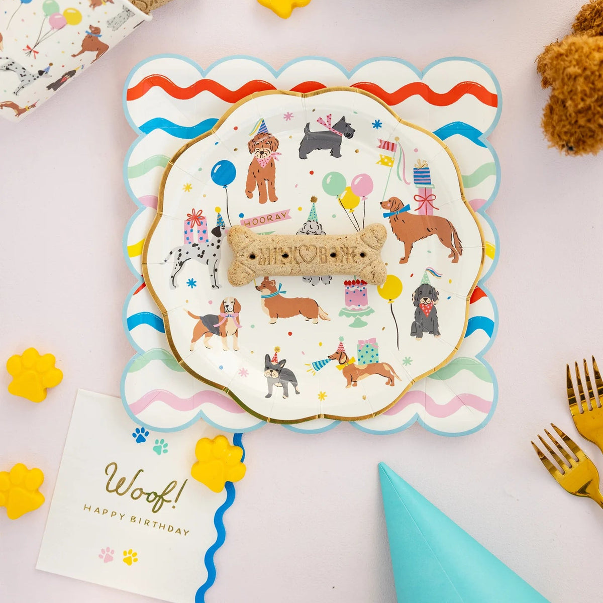 Puppy party flat lay with dog bone treats, party plates, and Woof napkin on the table.
