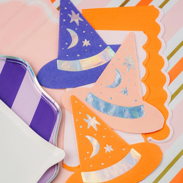 Trio of witch hat napkins in purple, peach, and orange styled next to striped plates with an orange scalloped guest towel peeking out beneath.