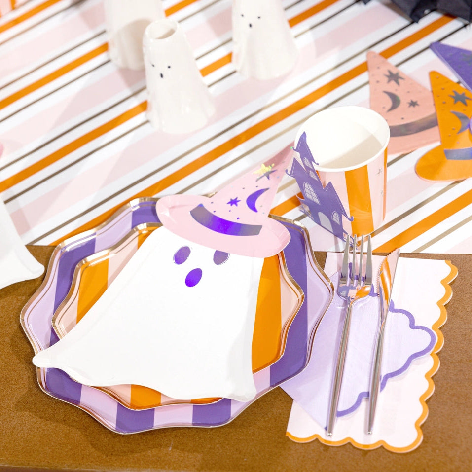Ghost plate wearing a pink hat layered over purple and orange striped plates with a lilac scalloped guest towel beside the cutlery and a haunted house cup.