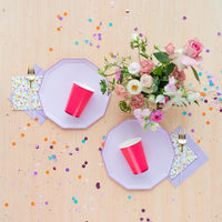 Minimal tablescape with two lilac plates, hot-pink cups, confetti, and florals