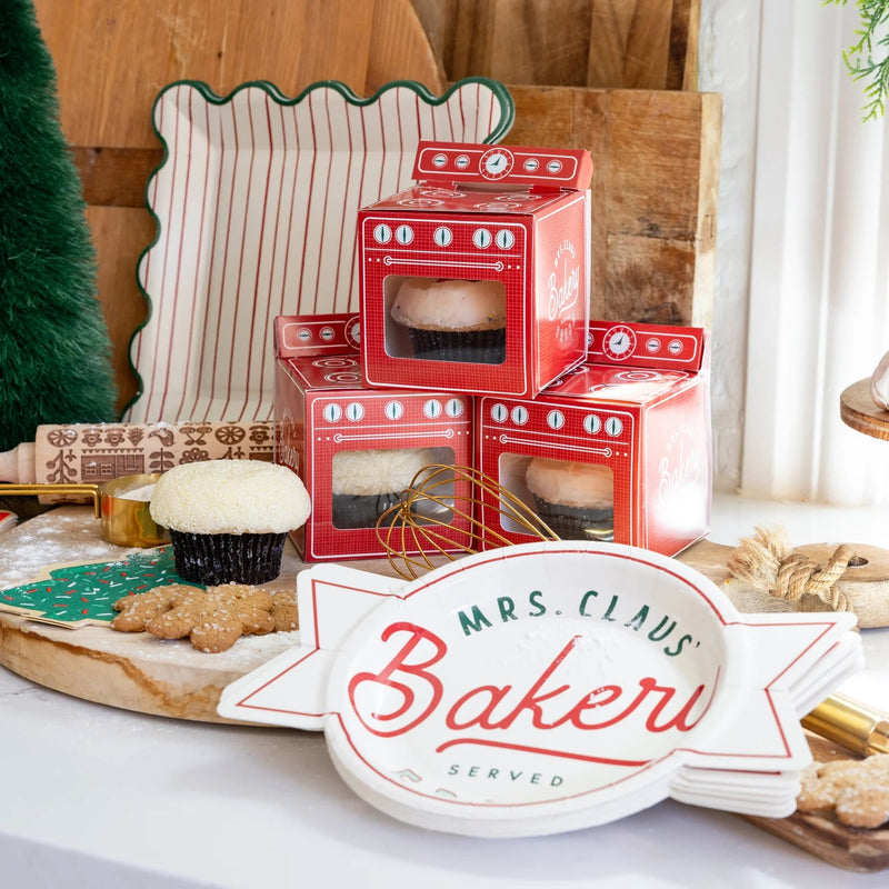 Holiday baking scene with stacked red oven treat boxes and “Mrs. Claus’ Bakery” holiday dessert plates on dessert table
