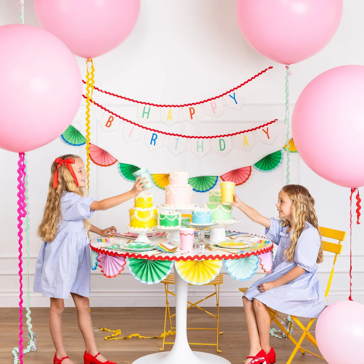 Two children toasting pastel party cups at a rainbow birthday table with bunting backdrop.