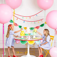 Two children toasting pastel party cups at a rainbow birthday table with bunting backdrop.